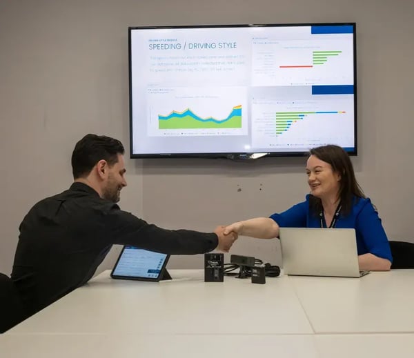 Transpoco employees in a meeting room shaking hands beneath a wall-mounted TV displaying telematics reports