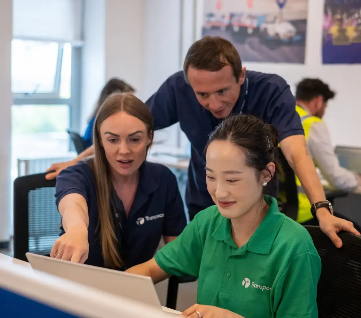 One Transpoco employee stands behind two others who are seated at a desk, with one pointing towards a screen