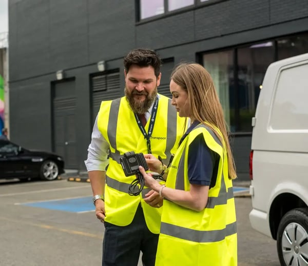 Two Transpoco employees in high-vis jackets standing next to a white van and reviewing a telematics device